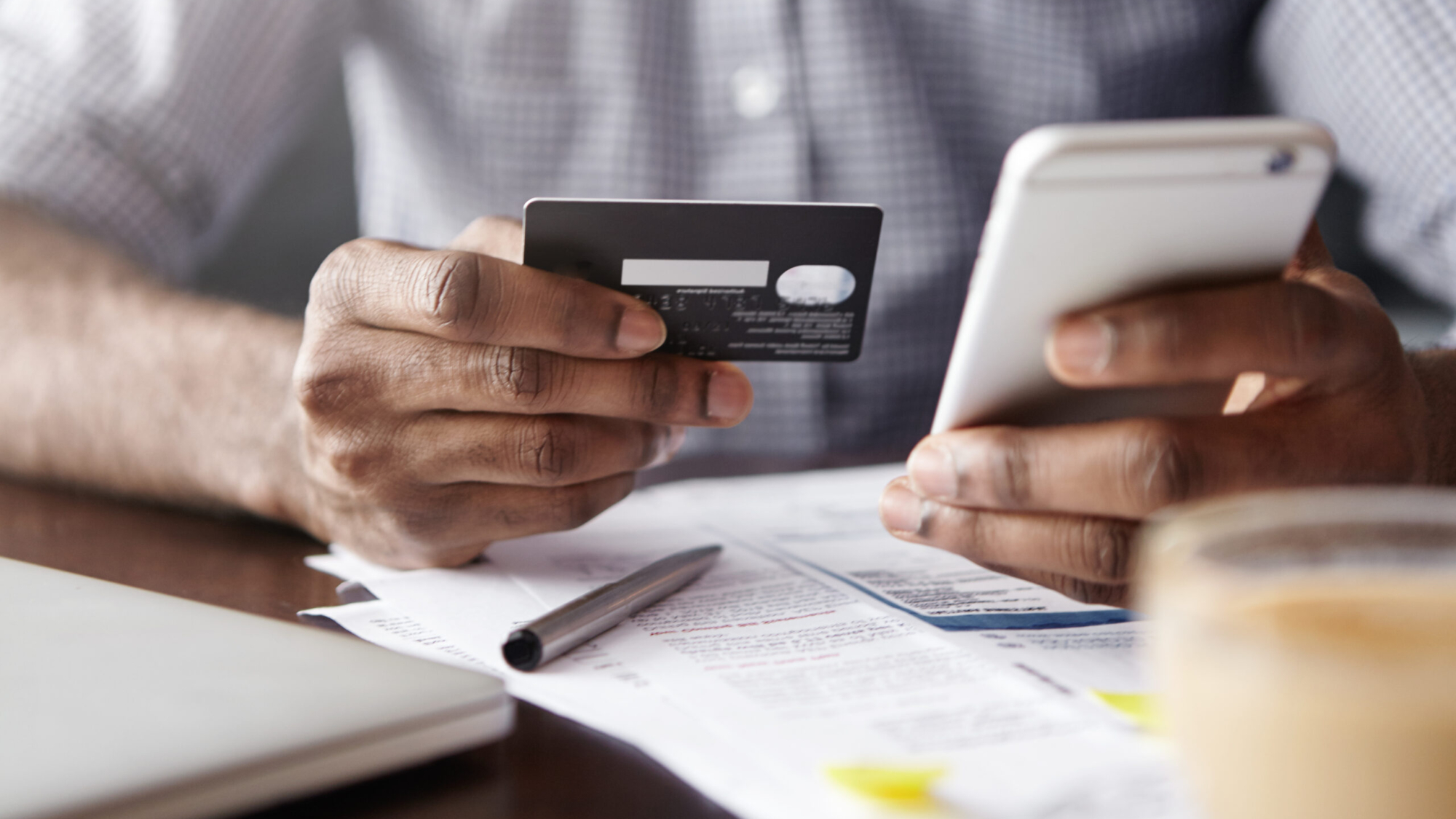 Close-up view of African man's hands holding plastic credit card and smart phone. Dark-skinned businessman using mobile banking application on his gadget while paying bills at cafe online via internet